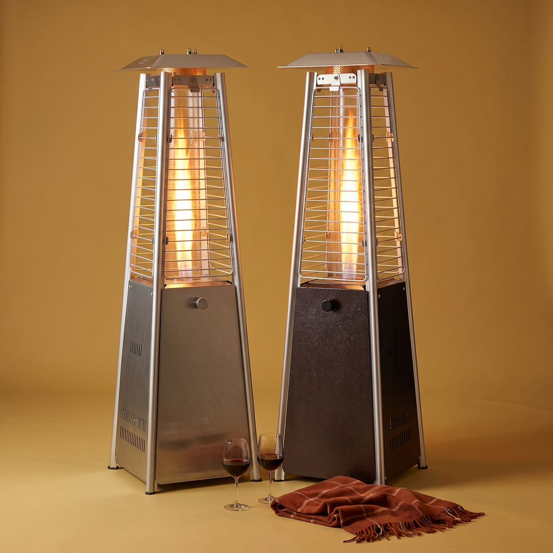 The Glass Flame tabletop glass tube patio heater in Silver and Hammered Black, lit, on a gold backdrop with wine glasses and a plaid throw