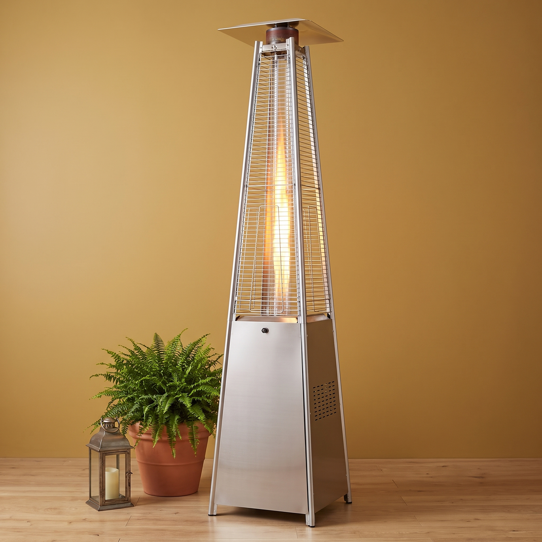 The Flame Tower pyramid propane patio heater in Gray glowing against a gold wall with a fern and lantern